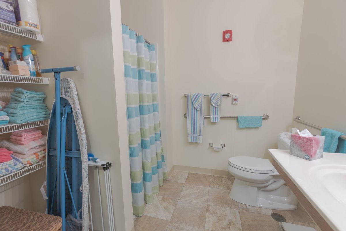 A bathroom with a shower curtain, toilet, sink, towels, and an ironing board.