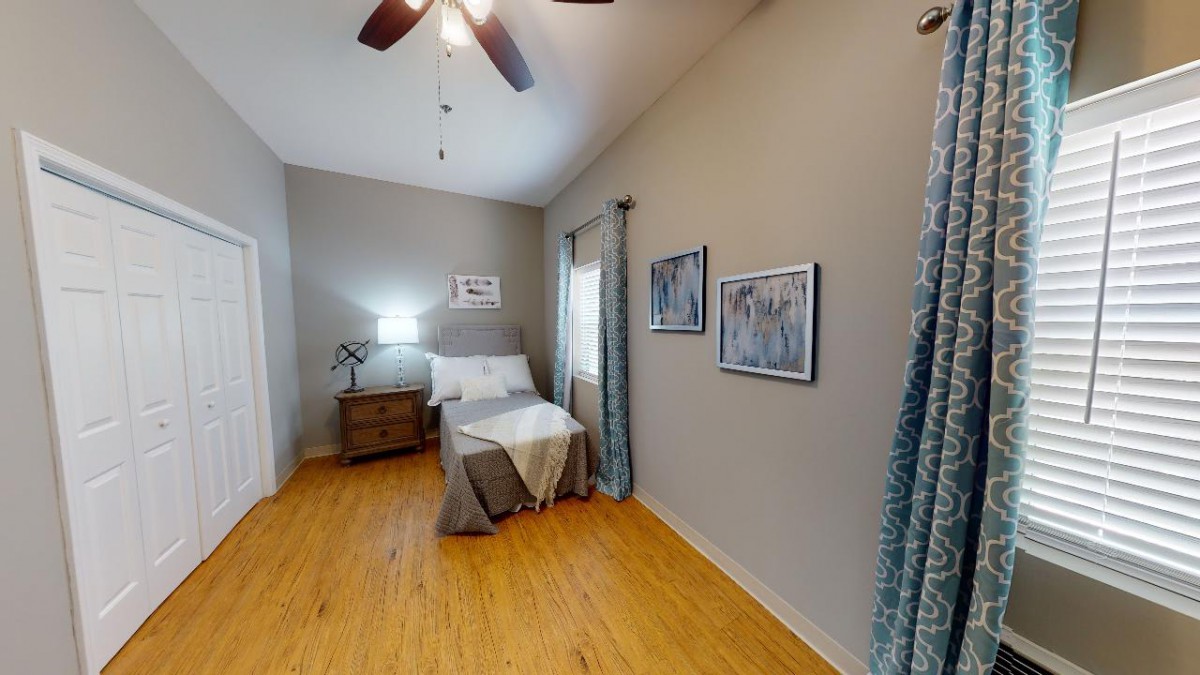 A cozy bedroom with a neatly made bed, nightstand, and lamp, featuring hardwood floors and modern decor.