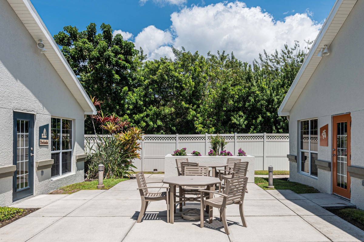 A sunny patio area with a table and chairs, surrounded by trees and a clear blue sky.