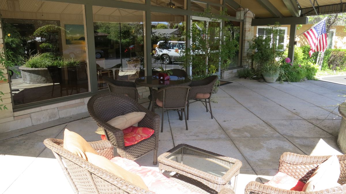 An outdoor patio area with wicker chairs and a table, surrounded by glass windows and greenery.