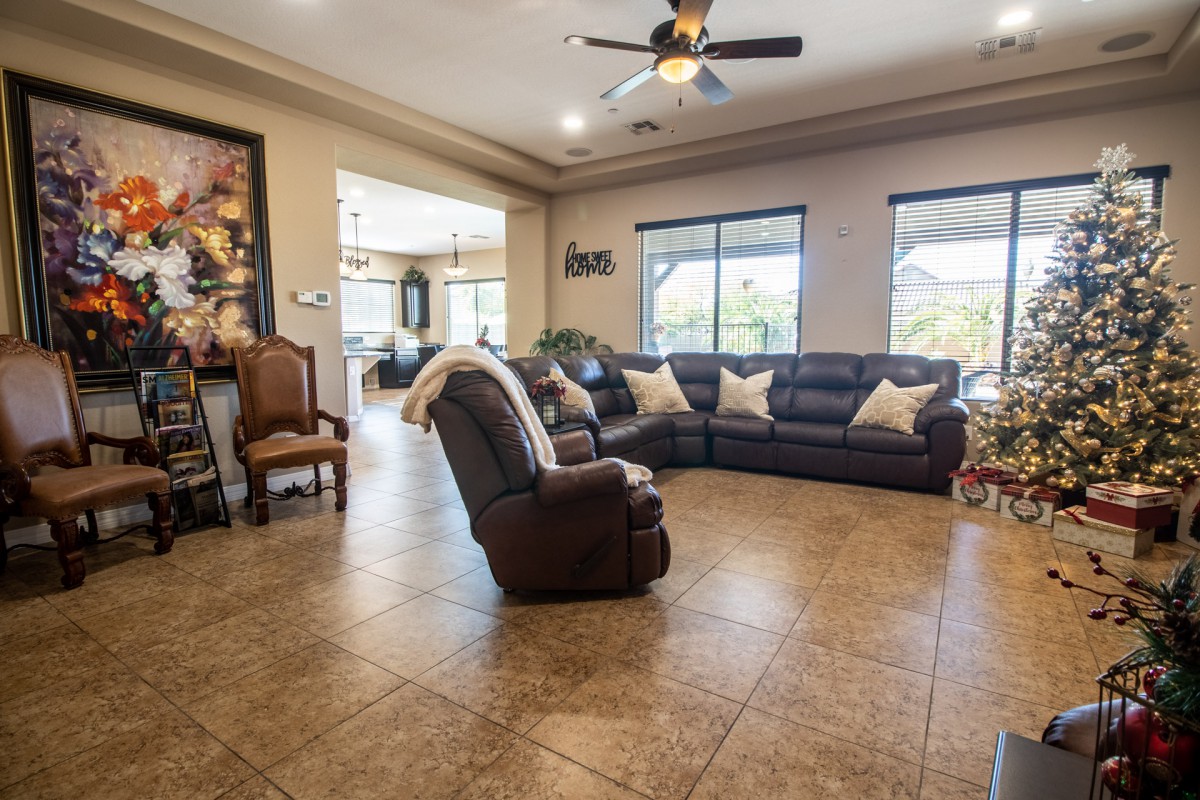 A spacious living room with a sectional sofa and armchair on a tiled floor, featuring large windows and a ceiling fan.