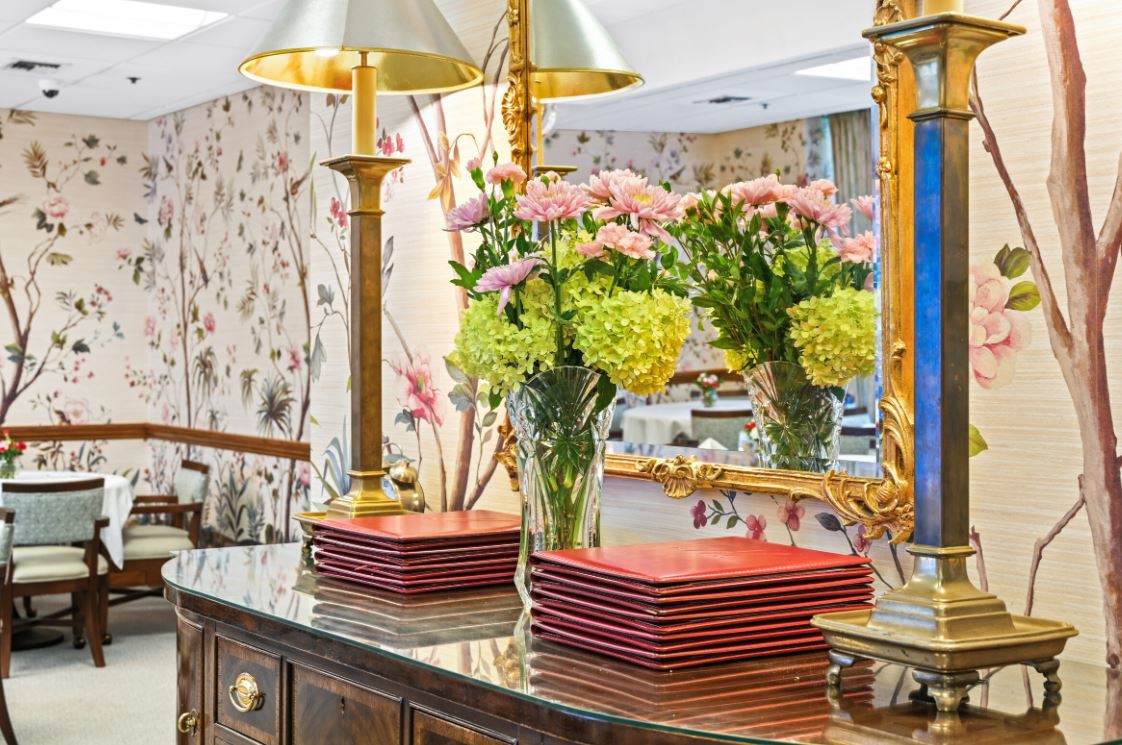 A sophisticated interior decor featuring floral arrangements in vases, gold lamps, and stacked plates on a polished wooden surface with a large ornate mirror.
