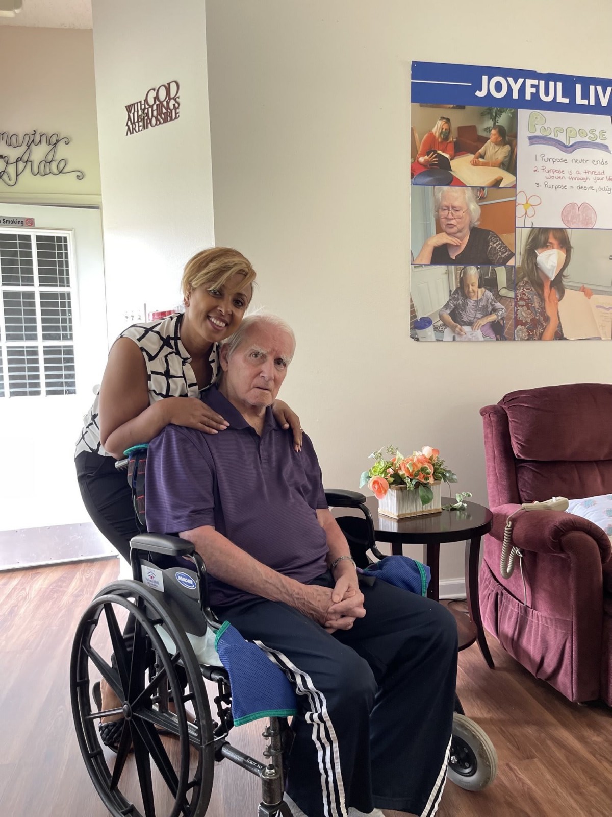 A caregiver smiling while standing behind an elderly person in a wheelchair, set in a cozy home environment.