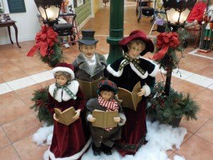 A display of Victorian-themed Christmas caroler dolls around a lamppost, set in a festive indoor scene with holiday decorations.