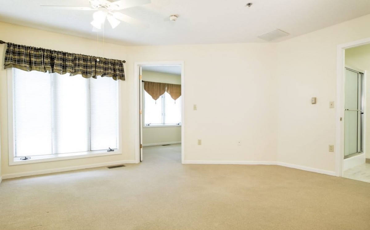 A spacious, empty room with carpet flooring, featuring large windows and a ceiling fan.