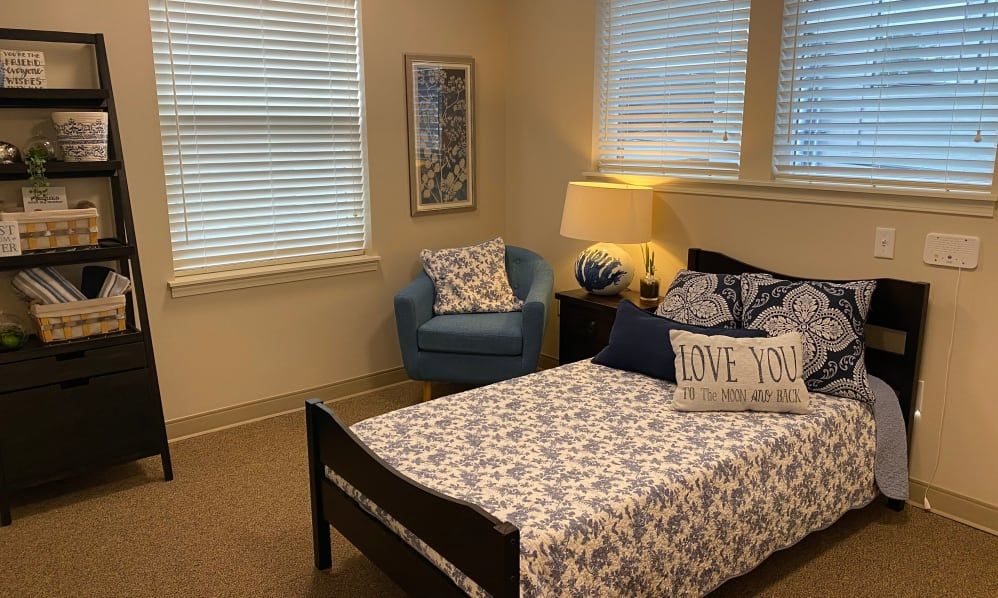A cozy bedroom featuring a neatly made bed with patterned bedding, a blue armchair, and a warmly lit lamp.