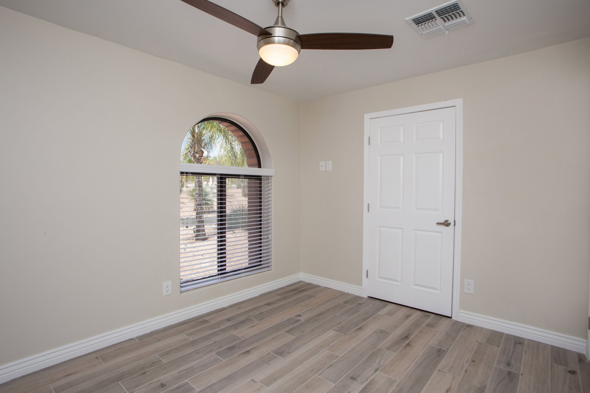 A modern empty room with a ceiling fan, arched window, and wood flooring.