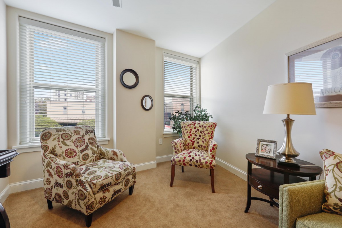 A cozy living room area with two patterned armchairs, a side table with a lamp, and large windows allowing natural light to fill the space.