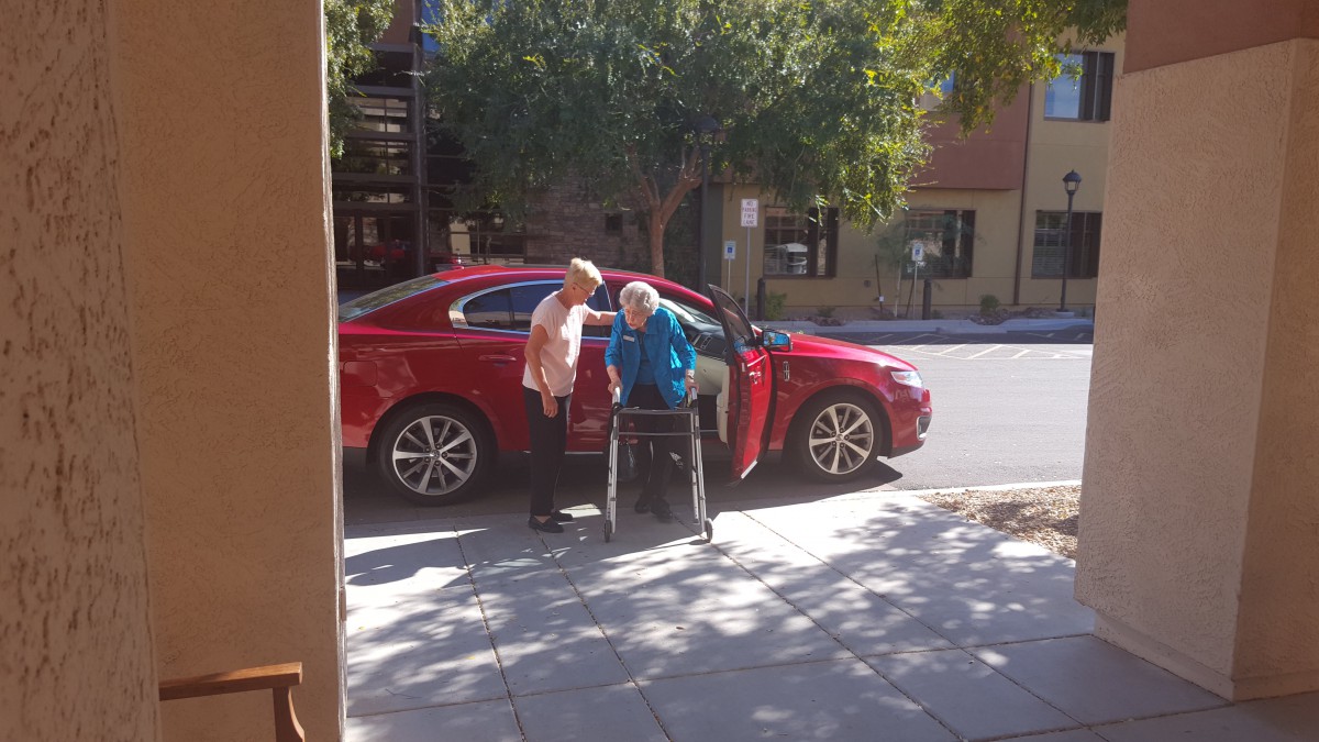 An elderly person using a walker receives assistance on a sunny street near a red car.