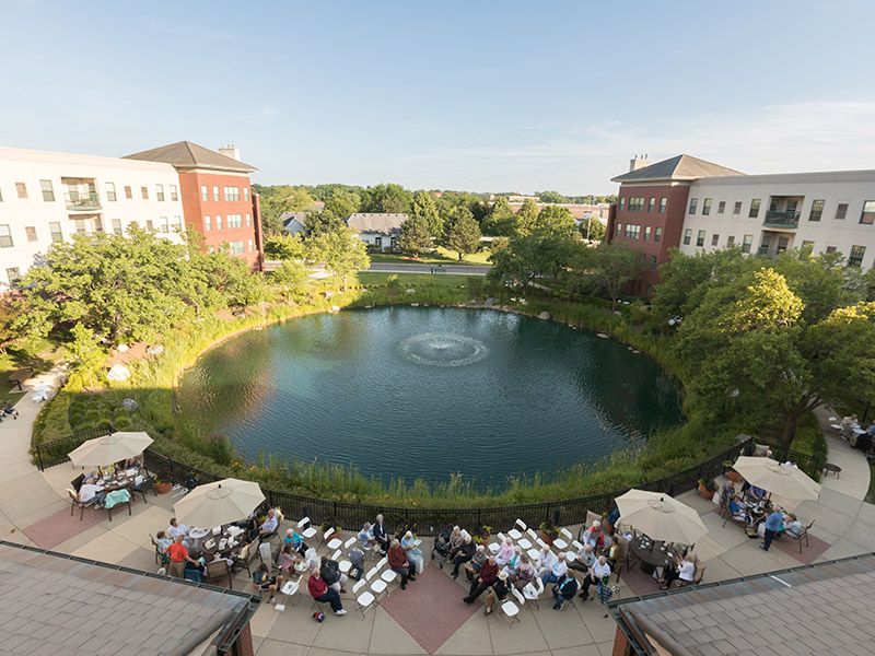 A scenic view of a pond surrounded by buildings, with tables and people gathered outdoors.