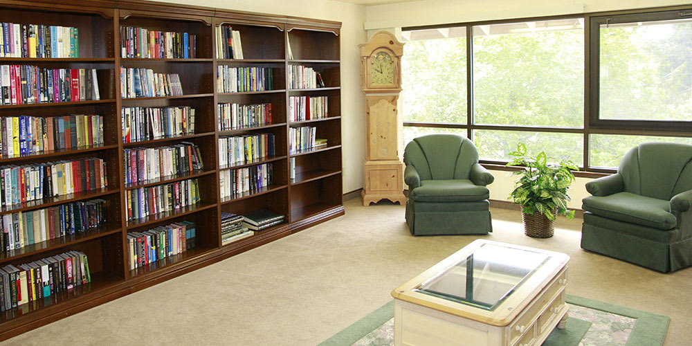 A cozy reading corner with a bookshelf, armchair, and grandfather clock by the window.