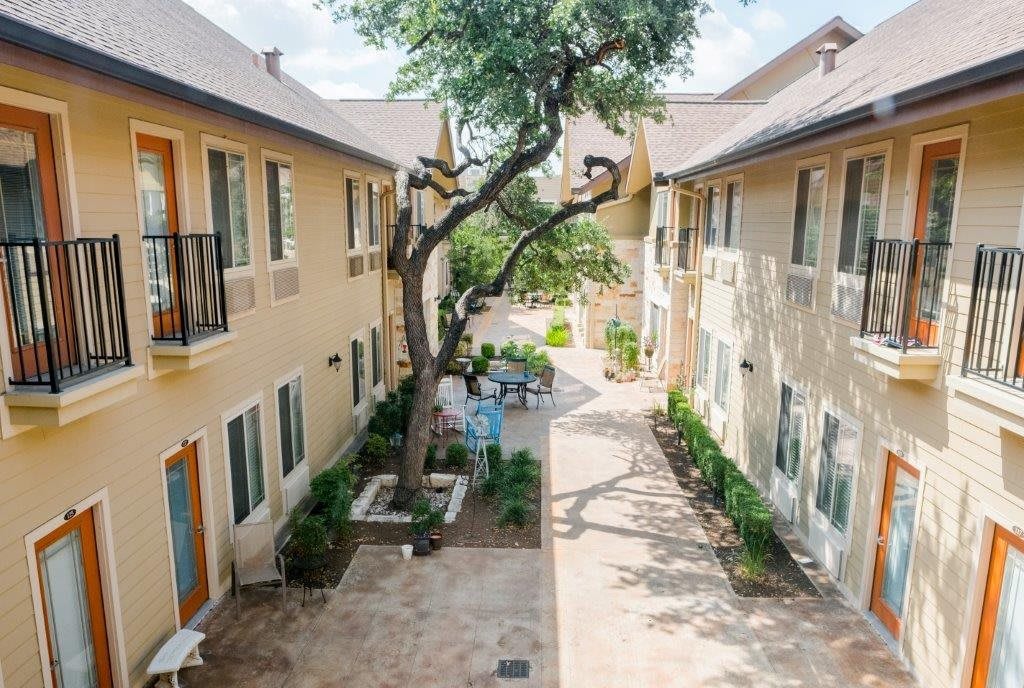 A serene courtyard between two apartment buildings with a central tree and outdoor seating.