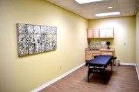 A clean and minimalistic treatment room with a massage table, decorated artwork on the yellow wall, and wooden flooring.