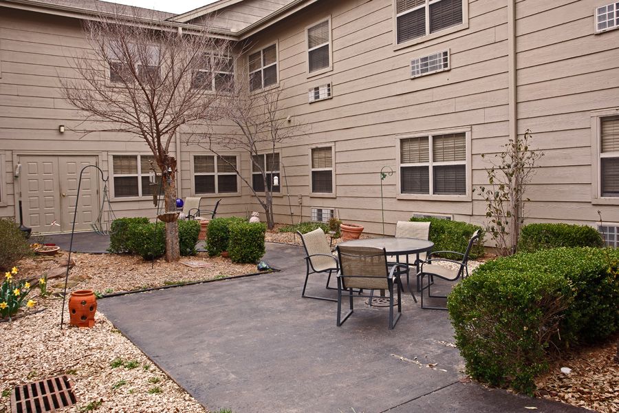 A cozy outdoor patio area with a table and chairs, surrounded by shrubs and adjacent to a building.