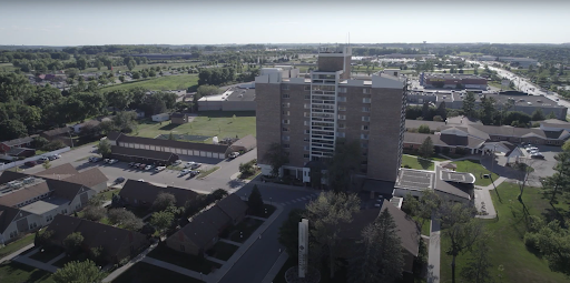 An aerial view of a multi-story building surrounded by green spaces and parking areas in an urban setting.