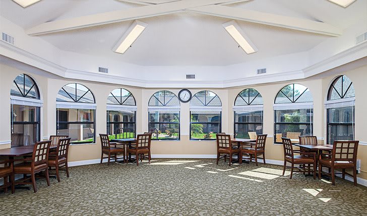 A bright room with multiple tables and chairs, large windows, and overhead lighting.