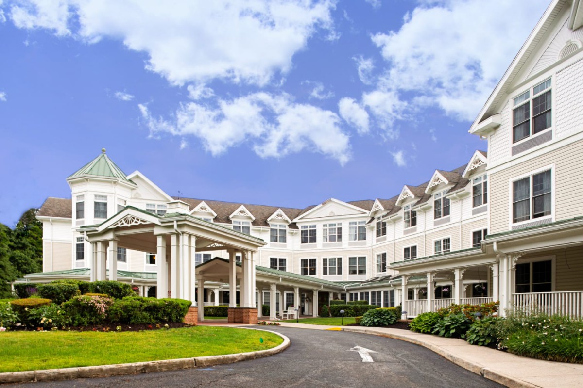 A large residential building with a covered entrance and surrounding greenery under a partly cloudy sky.