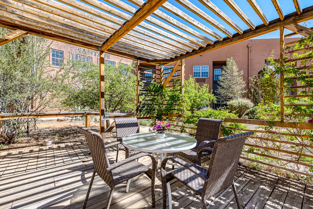A sunny outdoor patio area with a pergola, featuring a table and chairs surrounded by lush garden landscaping.