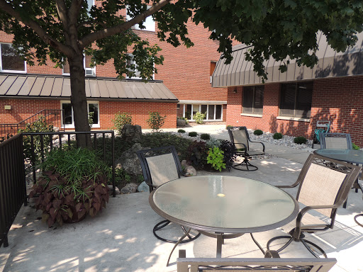 A serene outdoor patio area featuring a round glass table surrounded by chairs, complemented by lush greenery and a brick building backdrop.