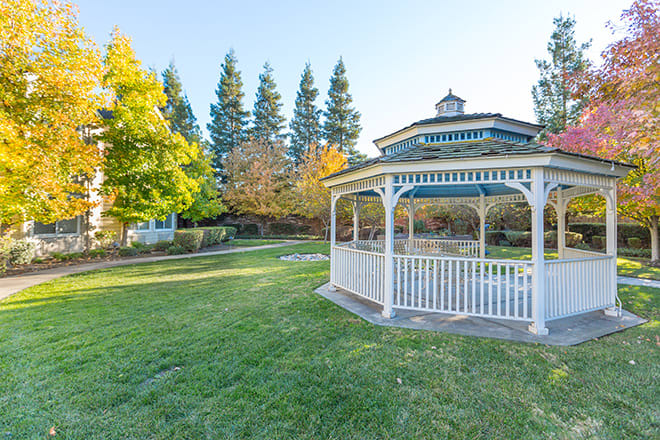 A scenic view of a park with a white gazebo, surrounded by grass and autumn trees.
