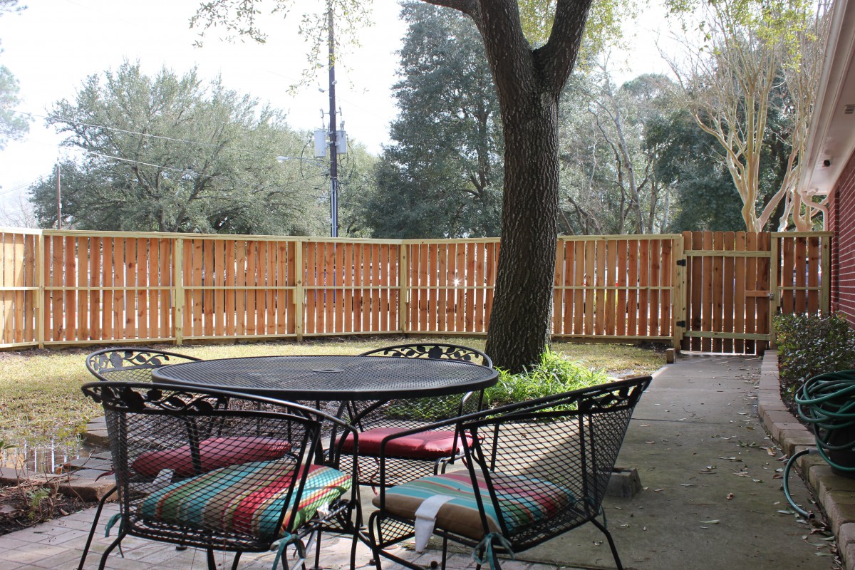 A cozy backyard patio with a metal table and chairs, accented by colorful cushions, under the shade of a large tree.