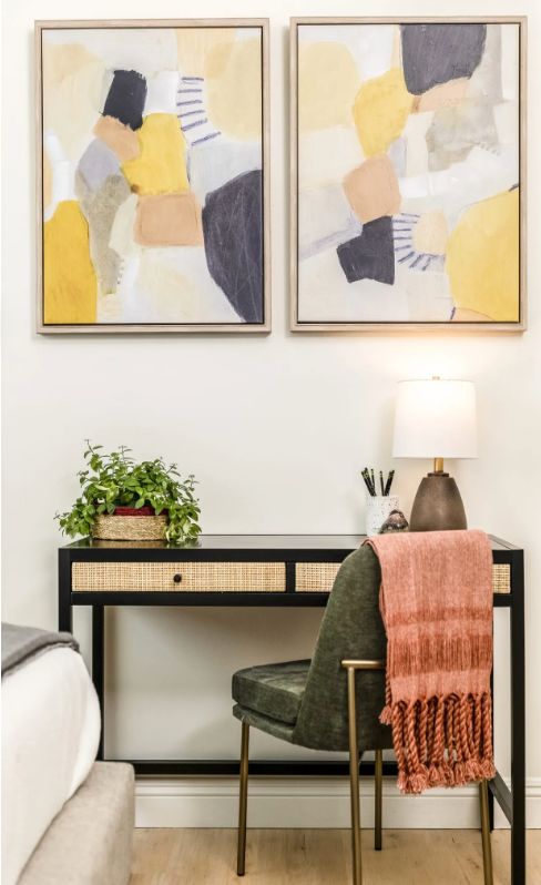 A stylish home office setup featuring a black desk with a woven front, a green chair draped with a pink throw, framed abstract art on the wall, and a potted plant next to a modern lamp.