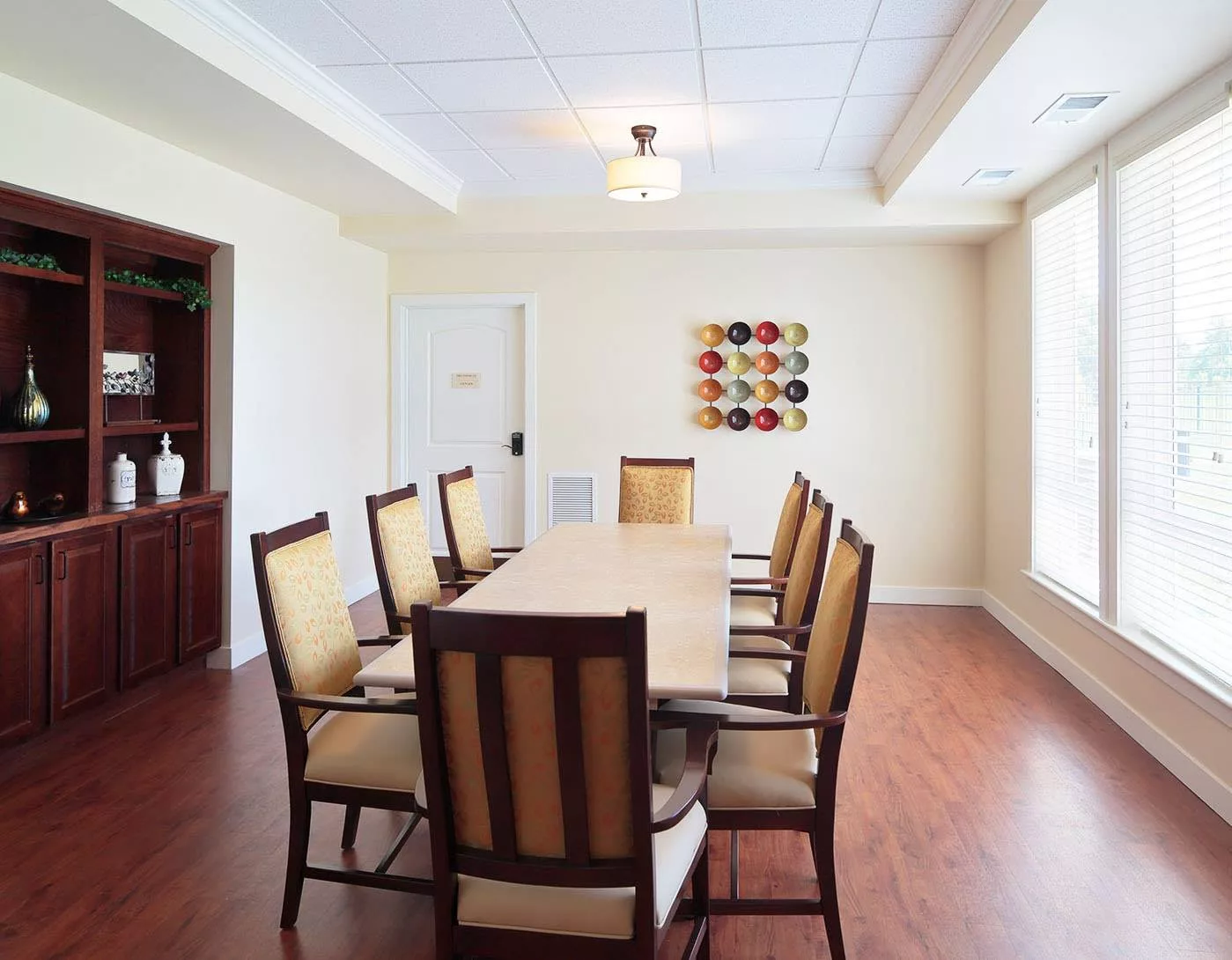 A well-lit dining room with a long table and several chairs, featuring wooden flooring and modern wall art.