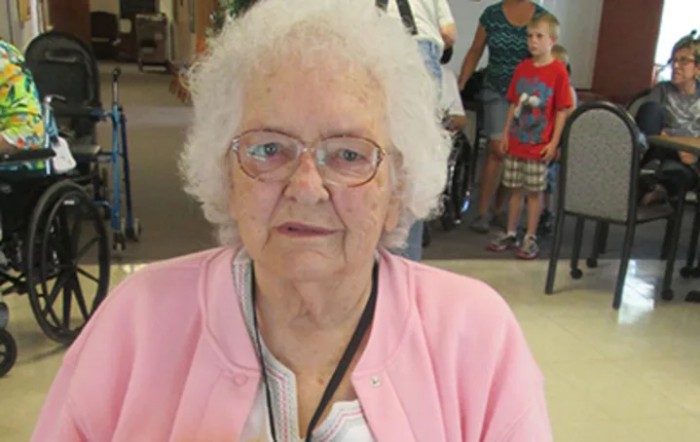 An elderly woman with white hair and glasses wearing a pink sweater indoors.