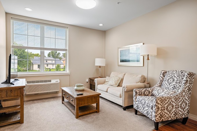 A cozy living room with a sofa, armchair, coffee table, and lamps, featuring a large window and wall art.