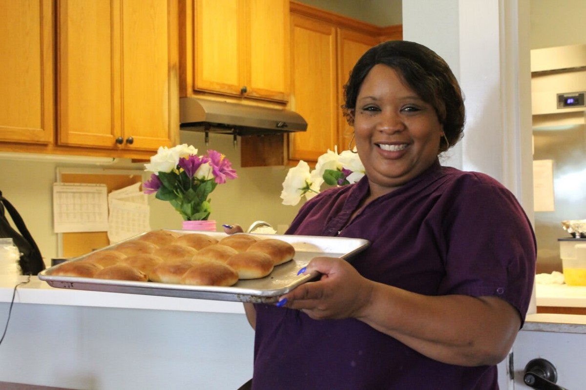 A woman smiling in a kitchen holding a tray of freshly baked bread with flowers in the background.