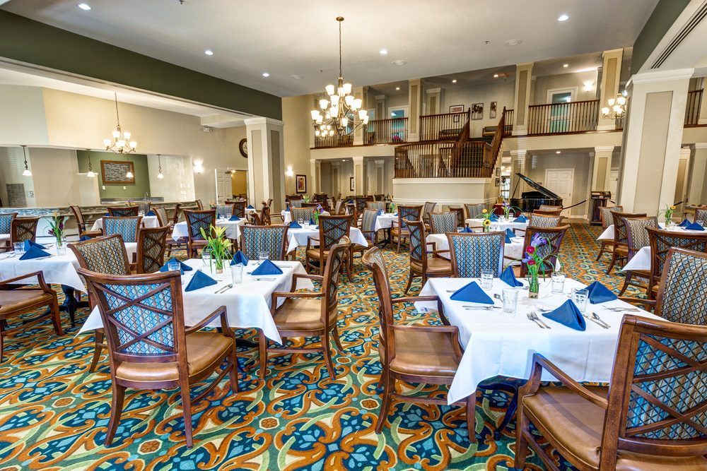 A spacious and elegantly decorated dining room with neatly arranged tables and chairs, featuring chandeliers and a vibrant carpet design.