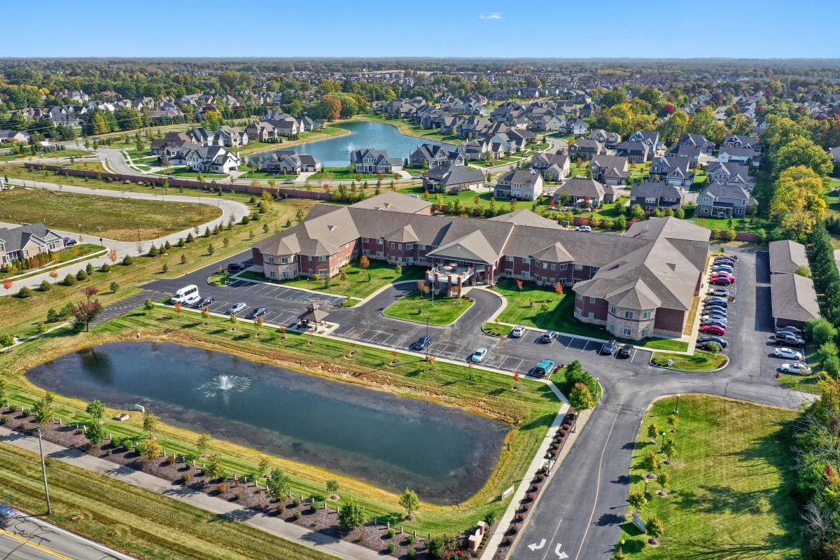 Aerial view of a suburban neighborhood featuring residential buildings, roads, and a small water body surrounded by greenery.