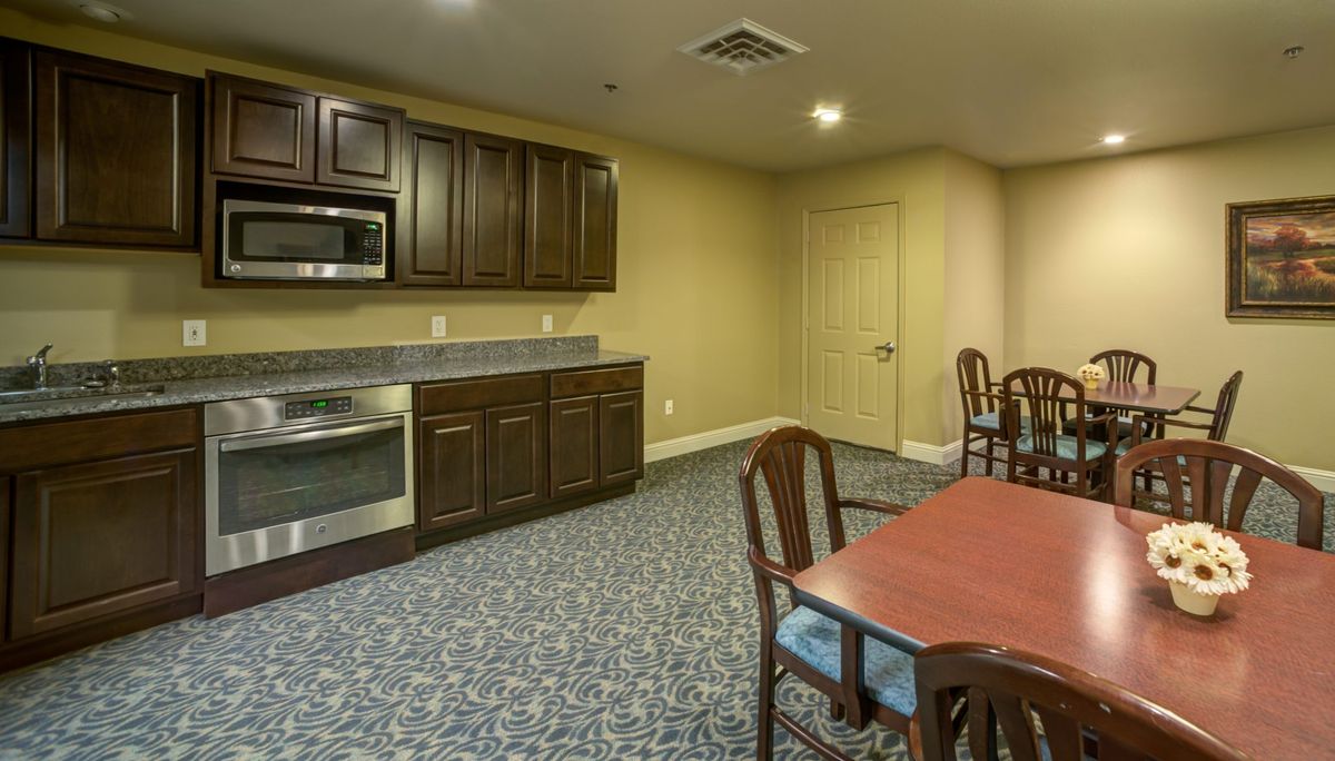 A view of a carpeted kitchen and dining area with wooden cabinets, a microwave, and a table with chairs.