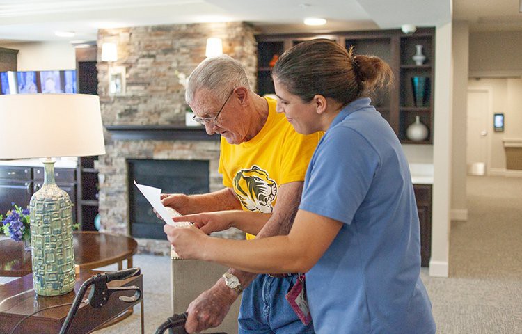 A caregiver assists an elderly man with reading a document inside a cozy living space.