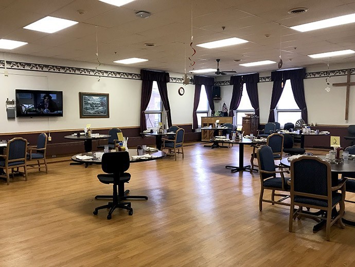An interior room with chairs and tables arranged in a spacious setting, featuring large windows with curtains.