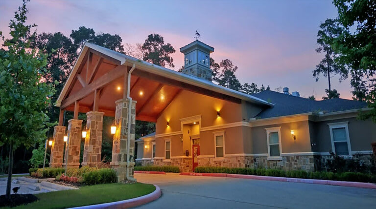 A warmly lit building entrance in the evening with a covered driveway and surrounding trees.