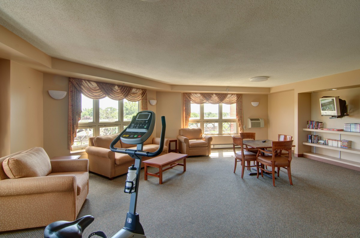 A spacious living room featuring an exercise bike, sofas, and a dining area with large windows allowing natural light.