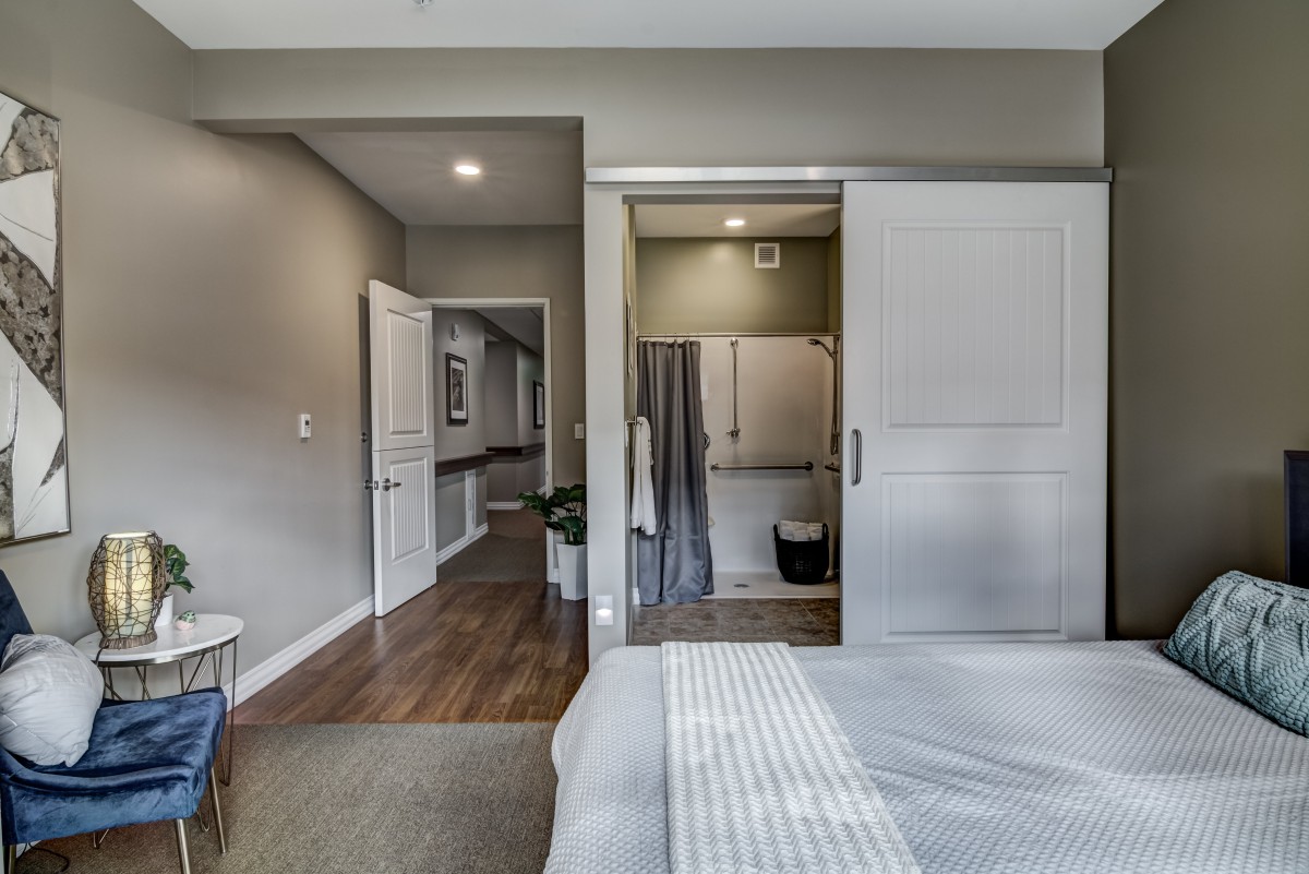 A modern bedroom with a view of an adjoining bathroom, featuring gray walls, a sliding door, and hardwood flooring.