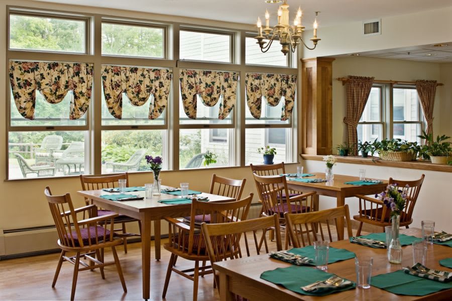 A cozy dining room with wooden furniture and natural light streaming through large windows, adorned with floral curtains.