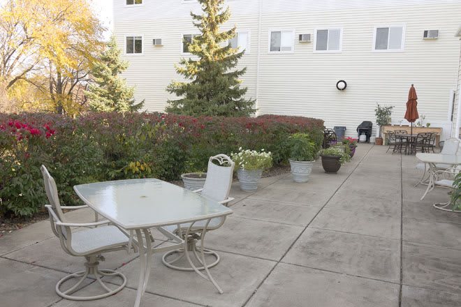 An outdoor patio area with a table and chairs, surrounded by plants and a building in the background.