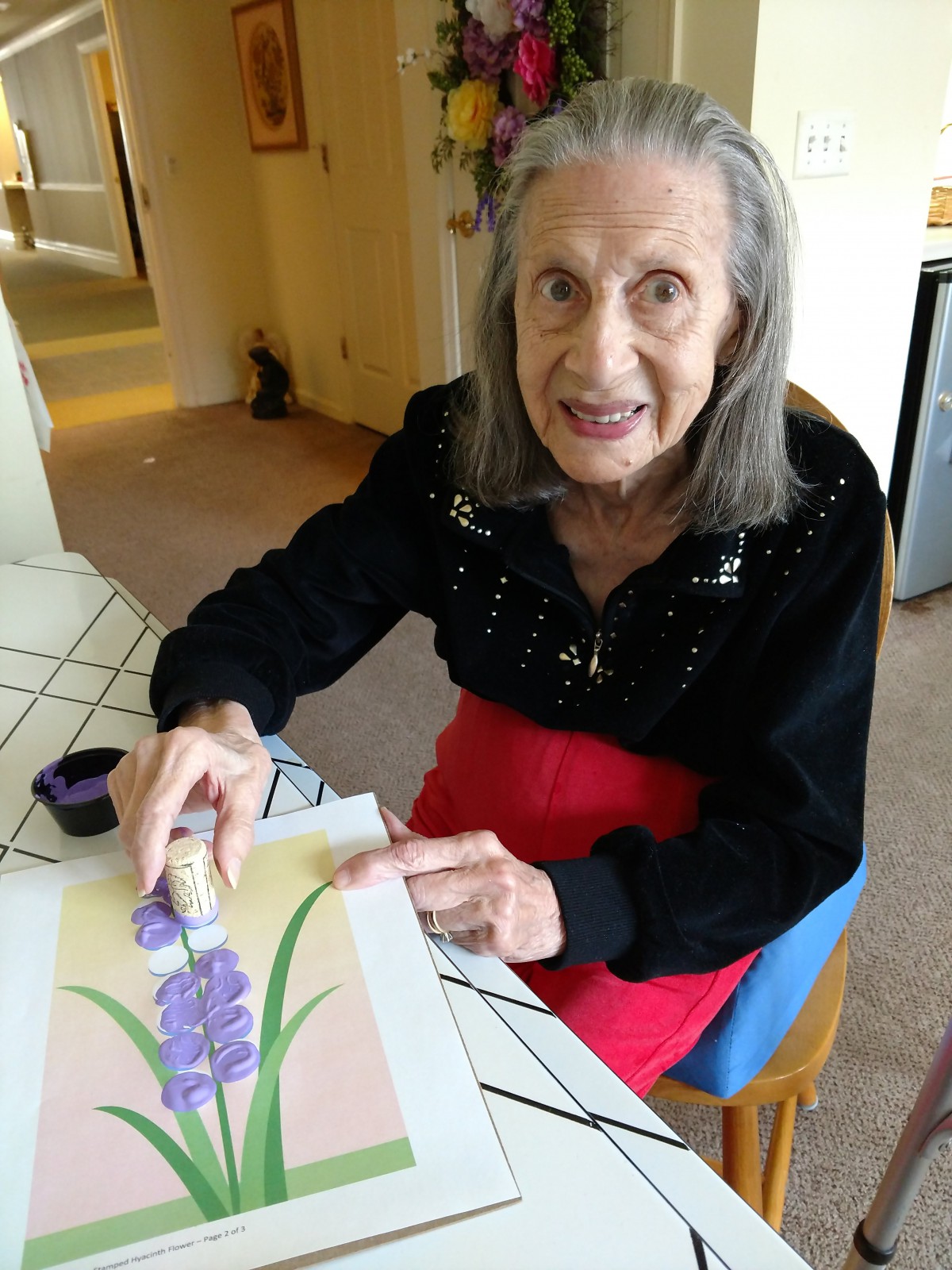 An elderly woman is sitting at a table engaged in painting a floral artwork.