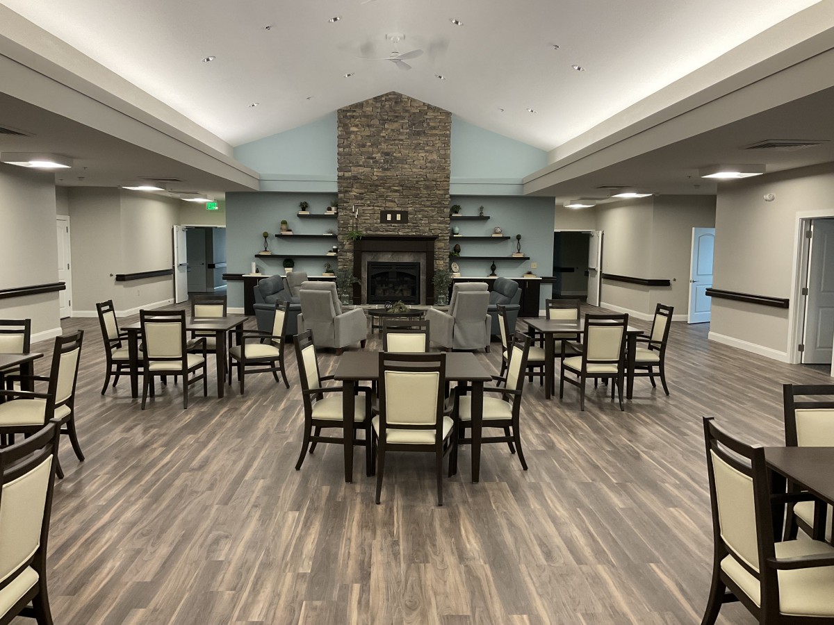 A spacious dining area with wooden flooring, featuring multiple tables and chairs, centered around a fireplace with surrounding shelves.