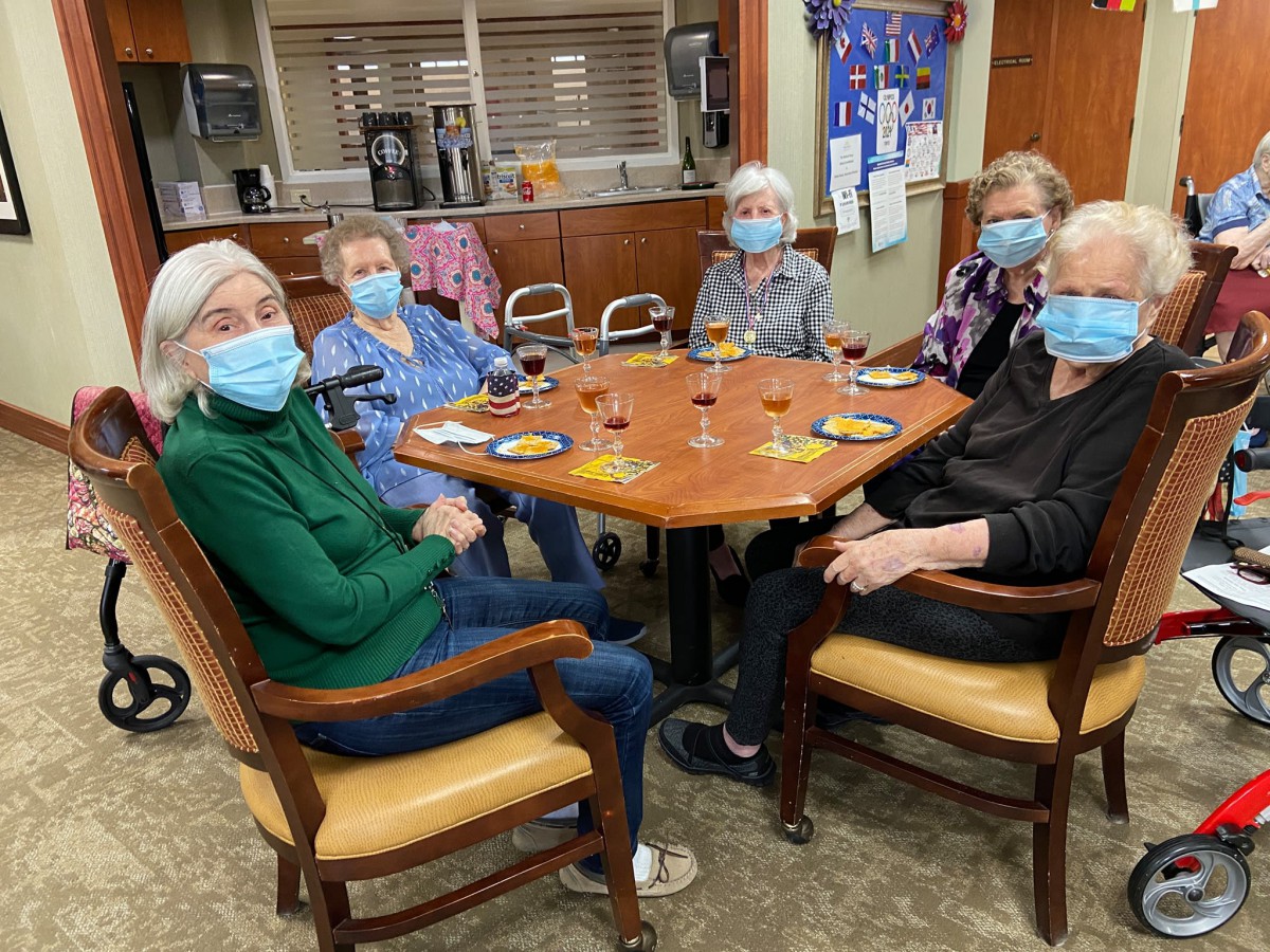 A group of elderly individuals wearing masks, sitting around a table and socializing indoors.