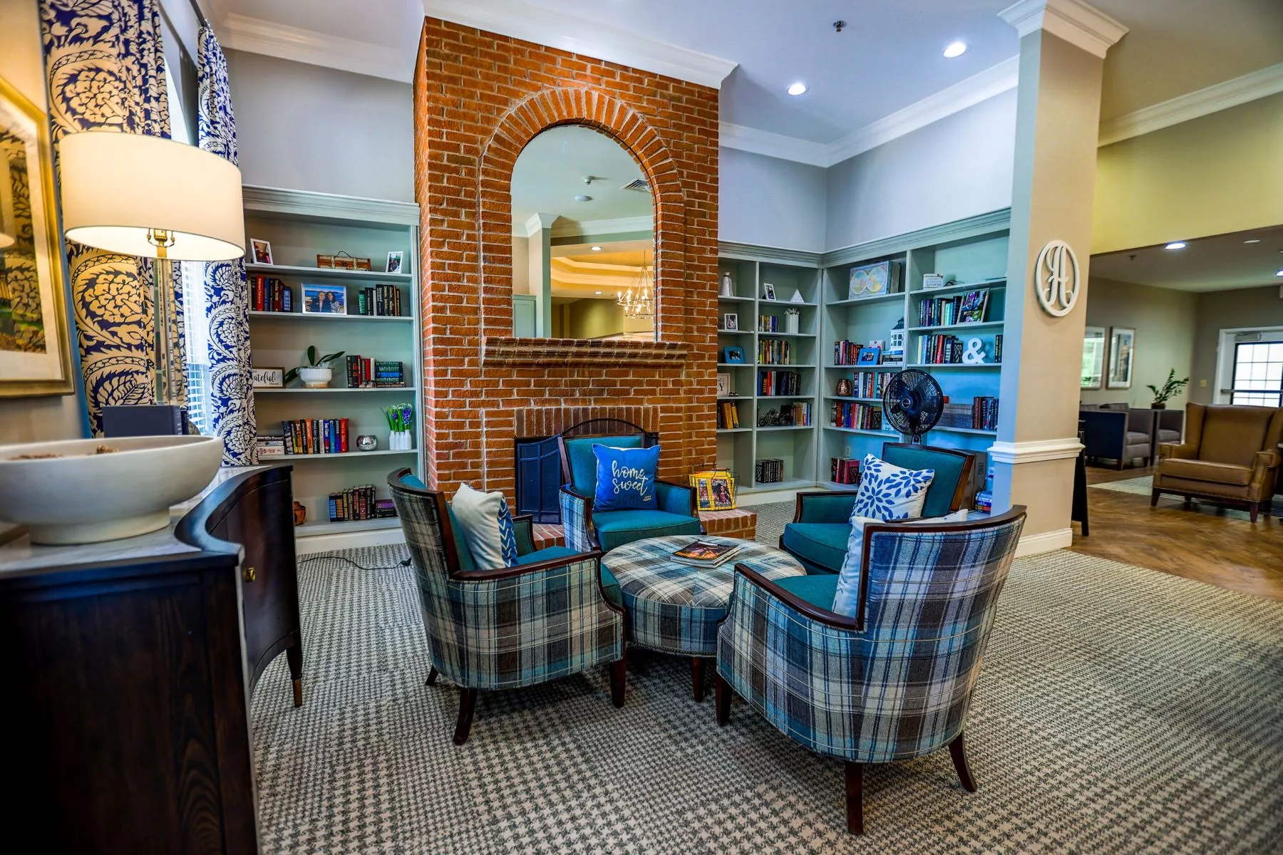 A cozy living room featuring a set of plaid armchairs arranged around a central ottoman, with a brick accent wall and bookshelves.
