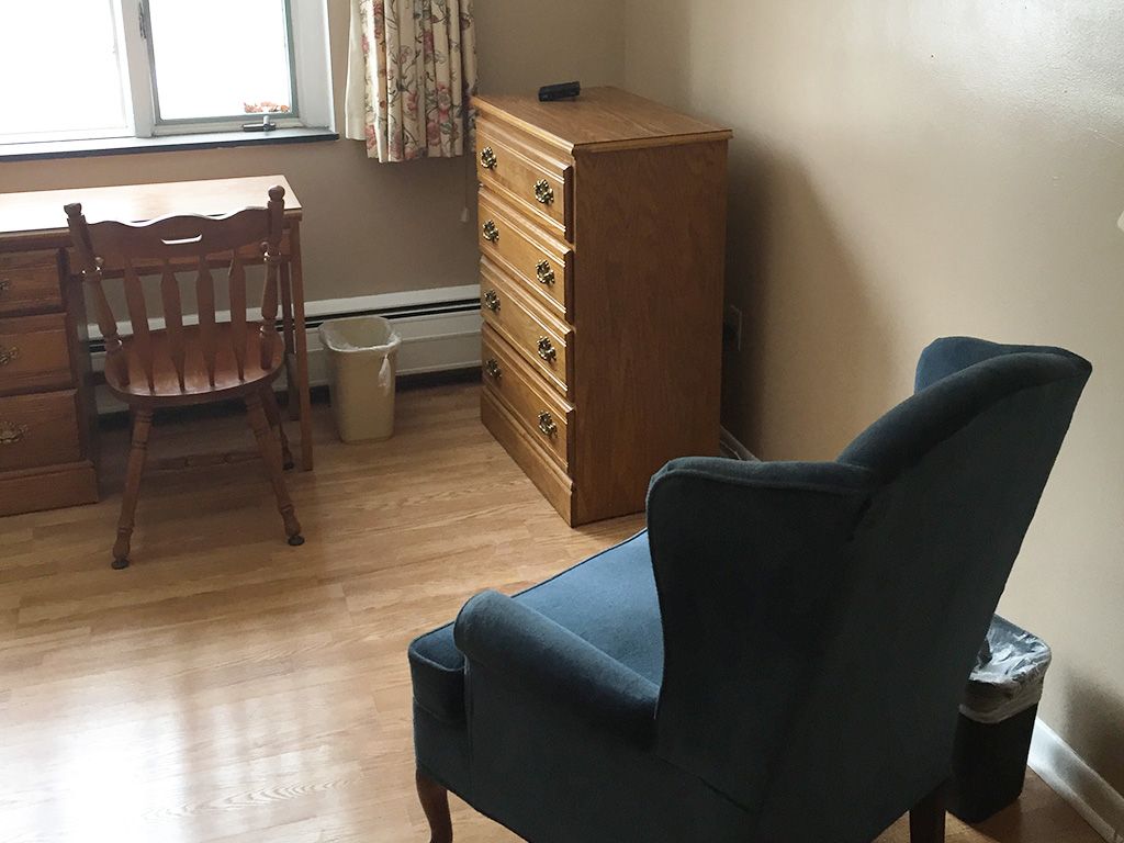 A simple room with a wooden desk, a blue armchair, and a dresser next to a window.