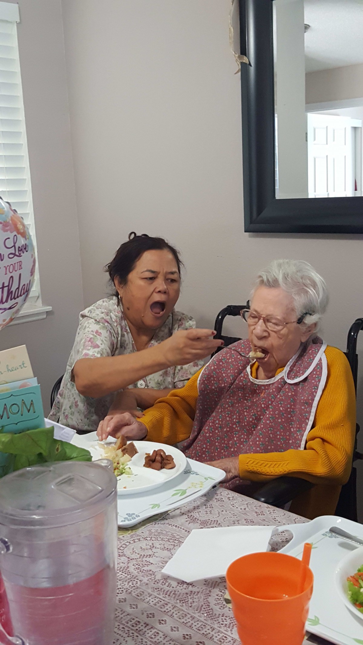 A caregiver feeds an elderly woman in a wheelchair during mealtime.