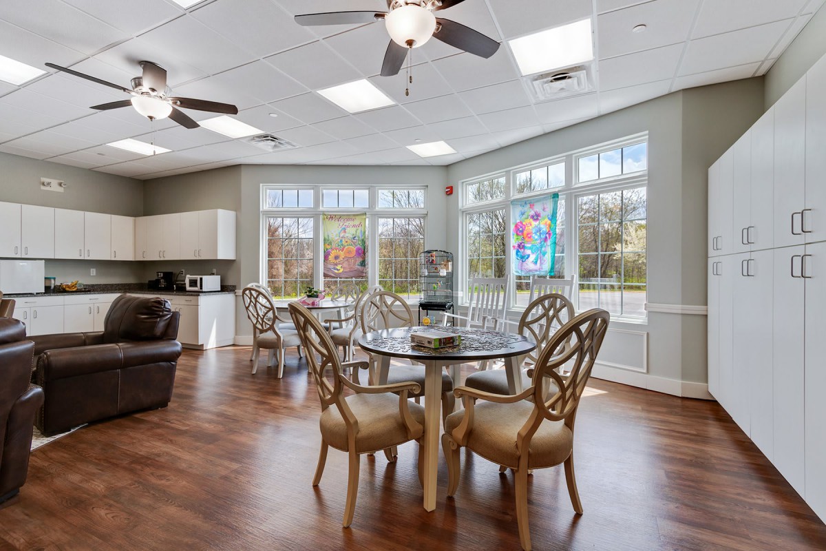 A modern dining area with ample natural light, featuring elegant tables and chairs, large windows, a kitchen counter, and ceiling fans.