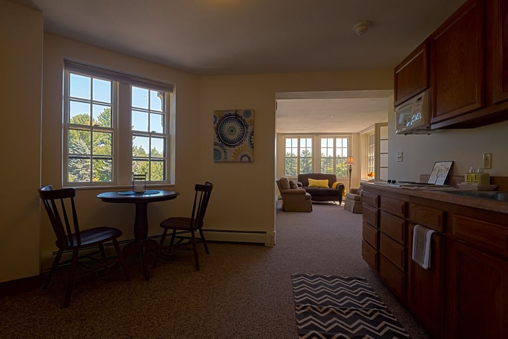 A cozy apartment living space with a kitchen, dining area, and a seating area filled with natural light from large windows.