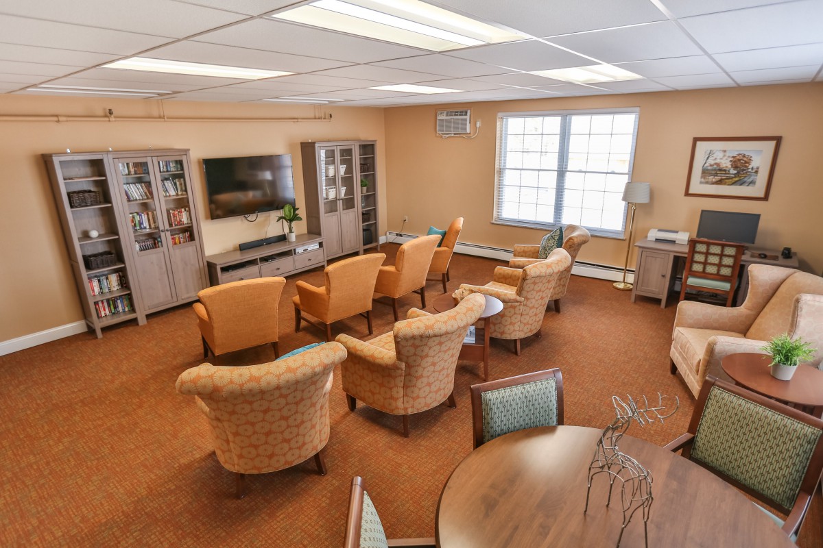 A cozy living room with multiple chairs arranged around a television, featuring carpeted flooring and large windows.