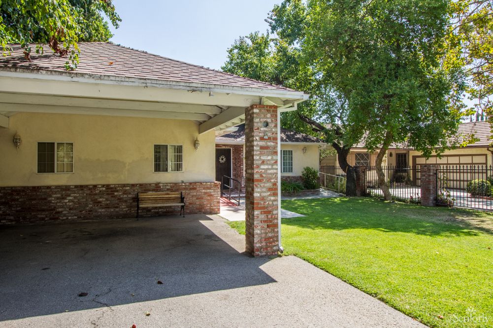 A sunny view of a house with a carport, surrounded by green lawn and trees.